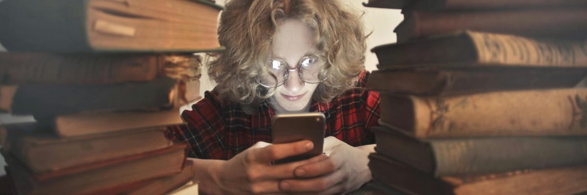 Young person wearing glasses looking at a phone sitting between tall stacks of books